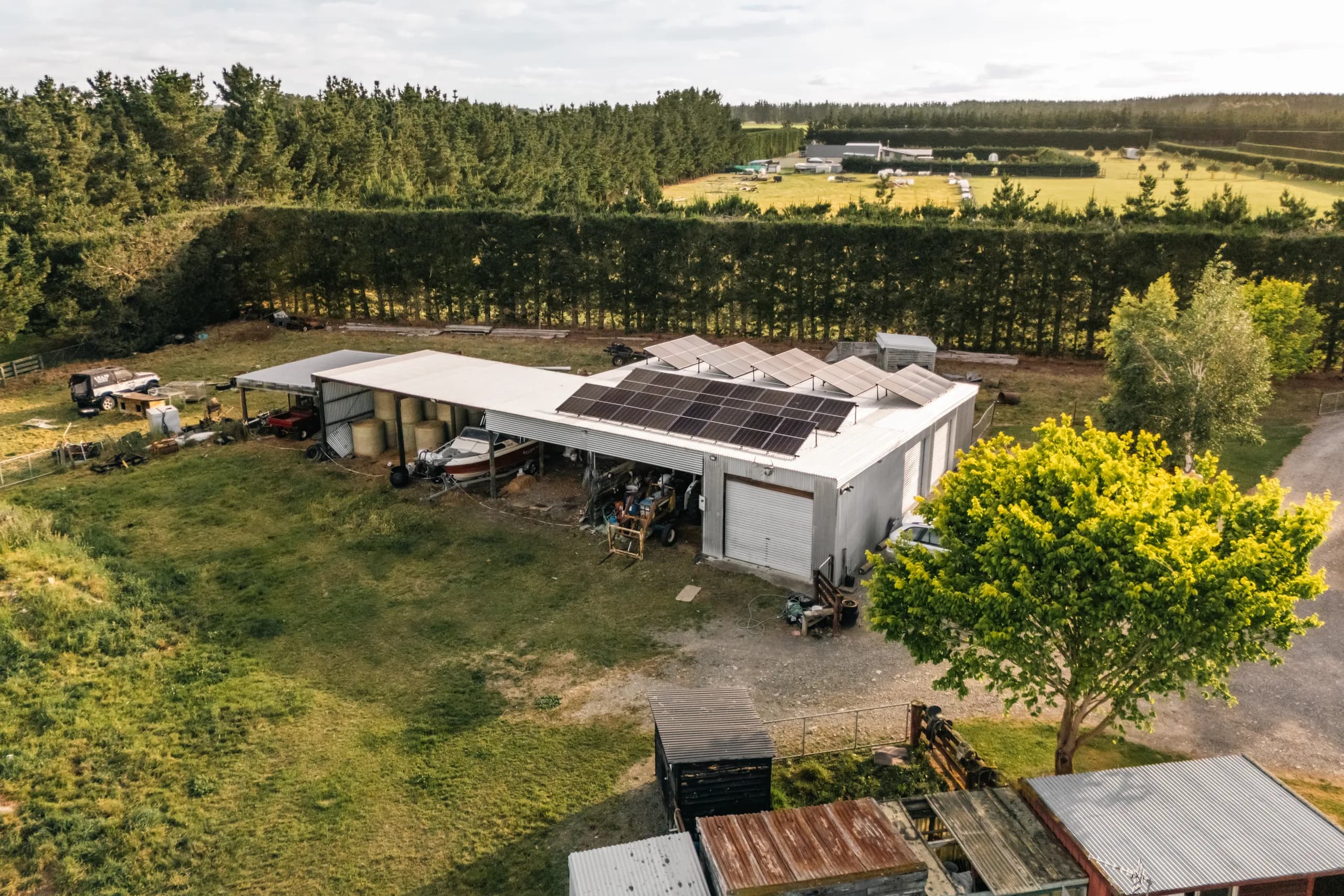Drone shot of a corrugated farm shed with a black solar panel array on its white roof, surrounded by a lifestyle-block yard with a boat, trailer and 4WD, tall hedges and green paddocks under a bright cloudy sky.