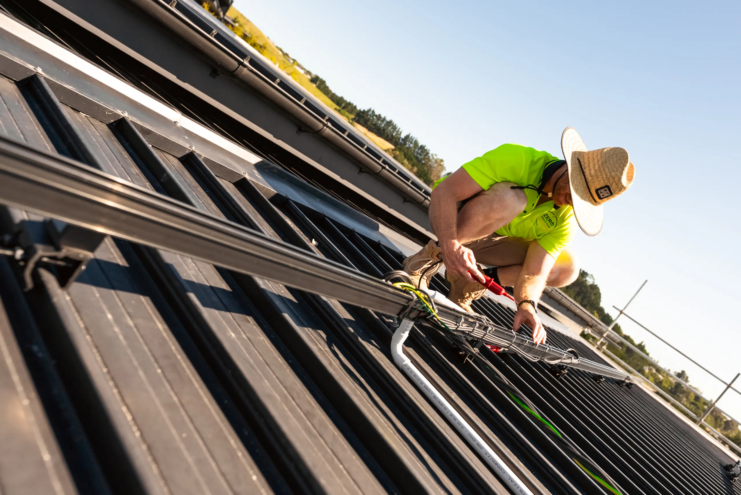 A Zero Electrical technician in a straw hat and hi-vis testing solar cabling with a multimeter on a dark metal roof.