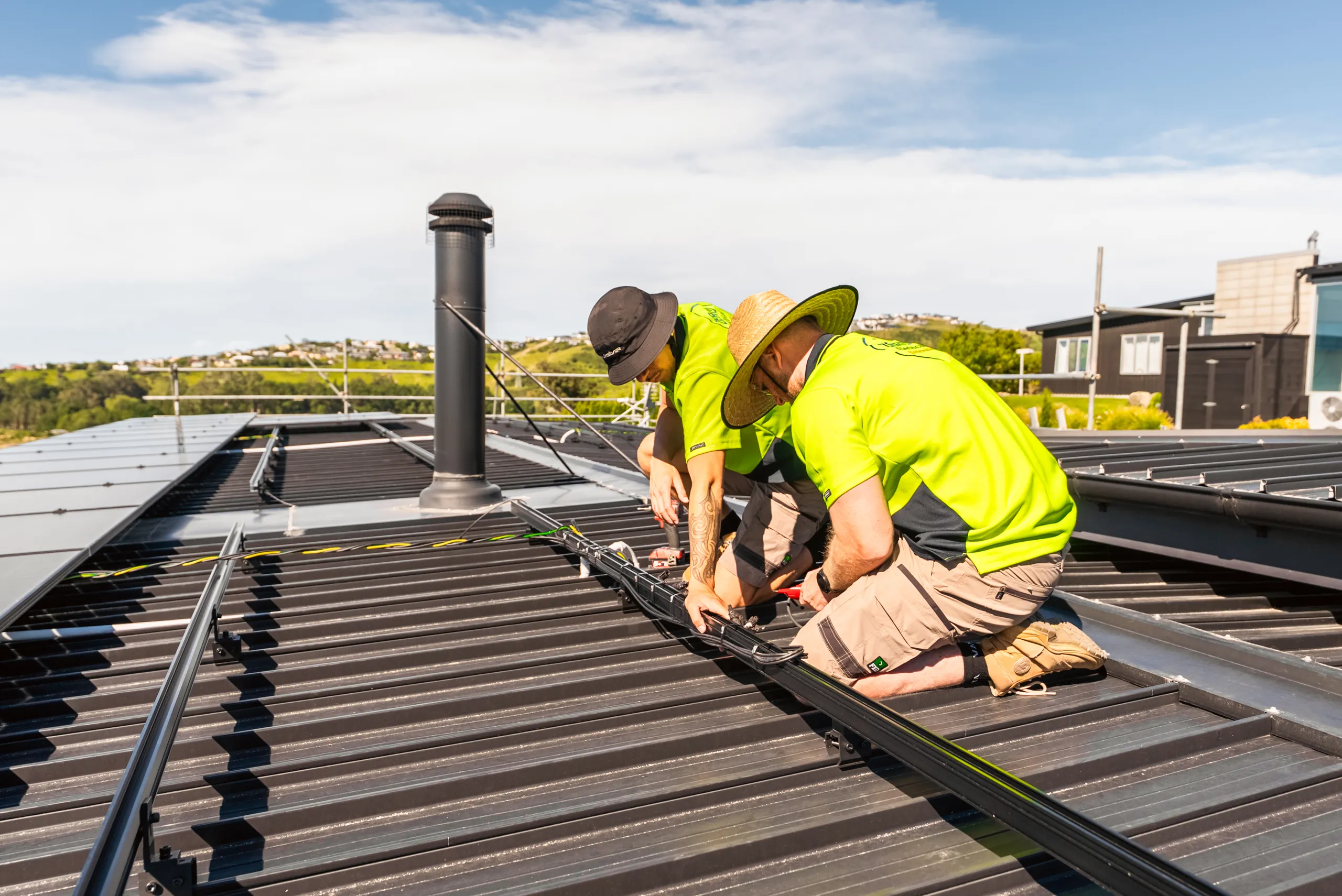 Two Zero technicians work on solar cabling beside a roof flue on a residential metal roof on a sunny day.