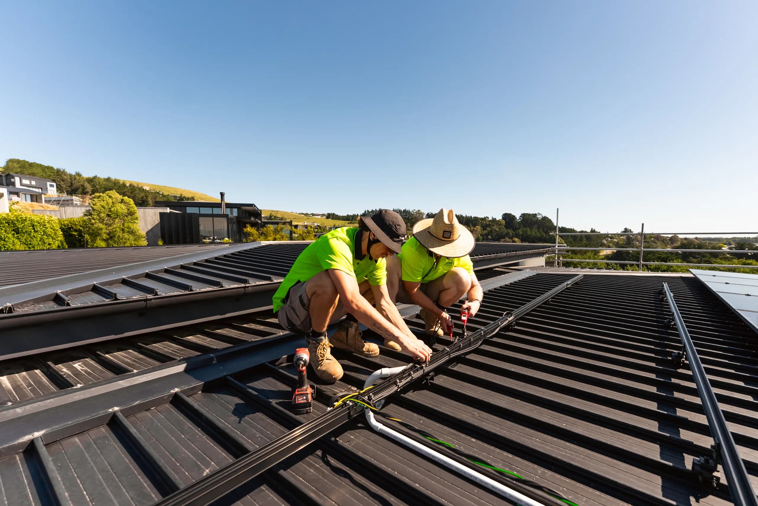 Two Zero technicians in hi-vis kneel to fit mounting rails and cabling for solar on a dark metal roof under blue sky.