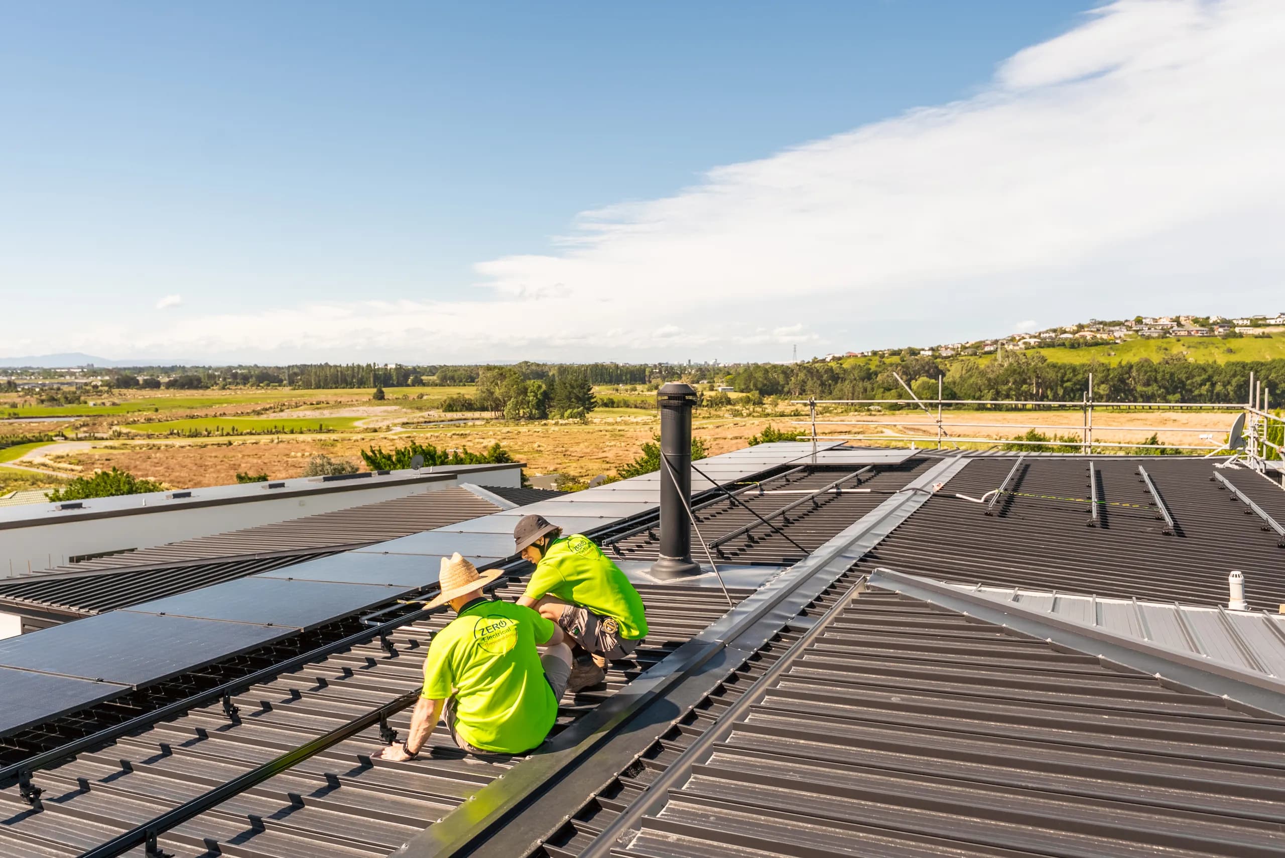 Two Zero technicians in branded hi-vis fit solar panels and rails on a large metal roof with a rural outlook.