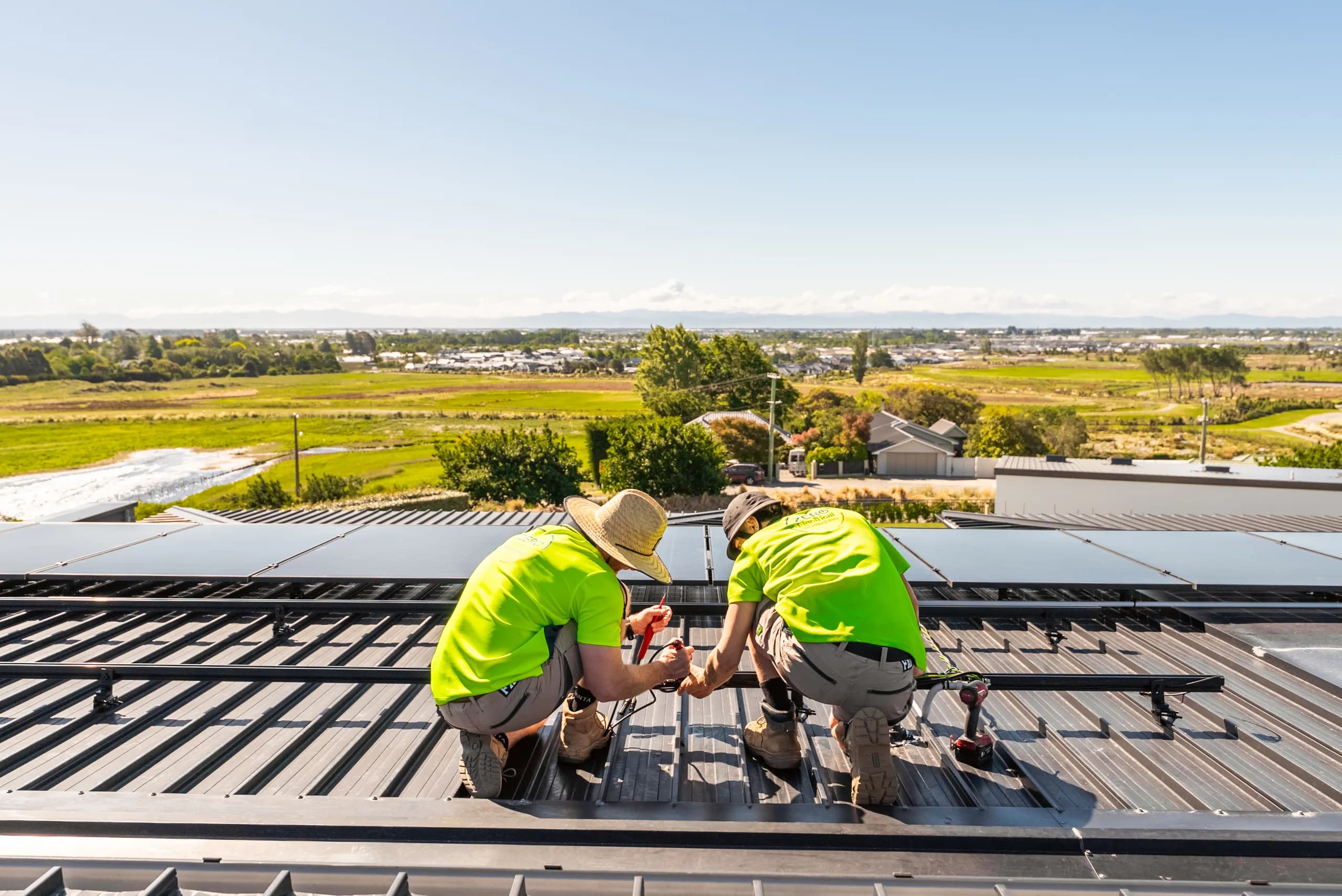 Two Zero technicians crouch to fit solar cabling on a rooftop overlooking green Canterbury paddocks and distant town.